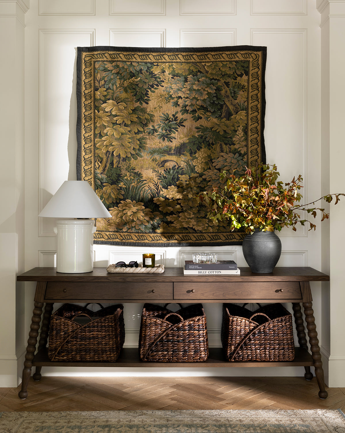 The Heathcliff Console, featuring a dark walnut oak finish and two drawers, sits against a white paneled wall topped by a green and gold tapestry. A white lamp, vase with branches, decor accents, and three woven baskets complete the look by McGee & Co.