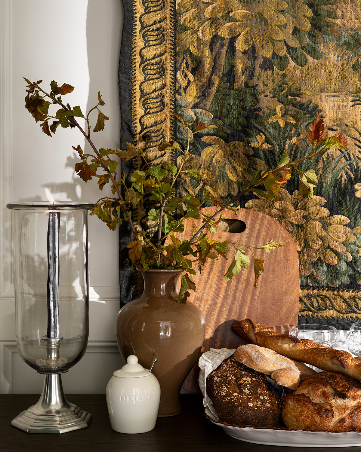 A decorative table setting features a lit candle in a glass holder, a brown vase with leafy branches, the Olive Stoneware Jar, and a plate of rustic breads, all arranged before a tapestry with green foliage patterns by McGee & Co.