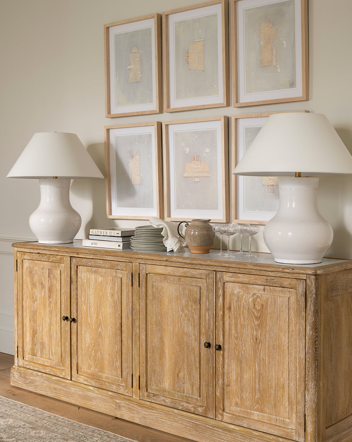 A wooden sideboard holds the Sorrento Table Lamp with a white linen shade, stacked books, dishes, an earthenware jug, and wine glasses. Above, six light-framed abstract artworks are displayed on the wall, by McGee & Co.