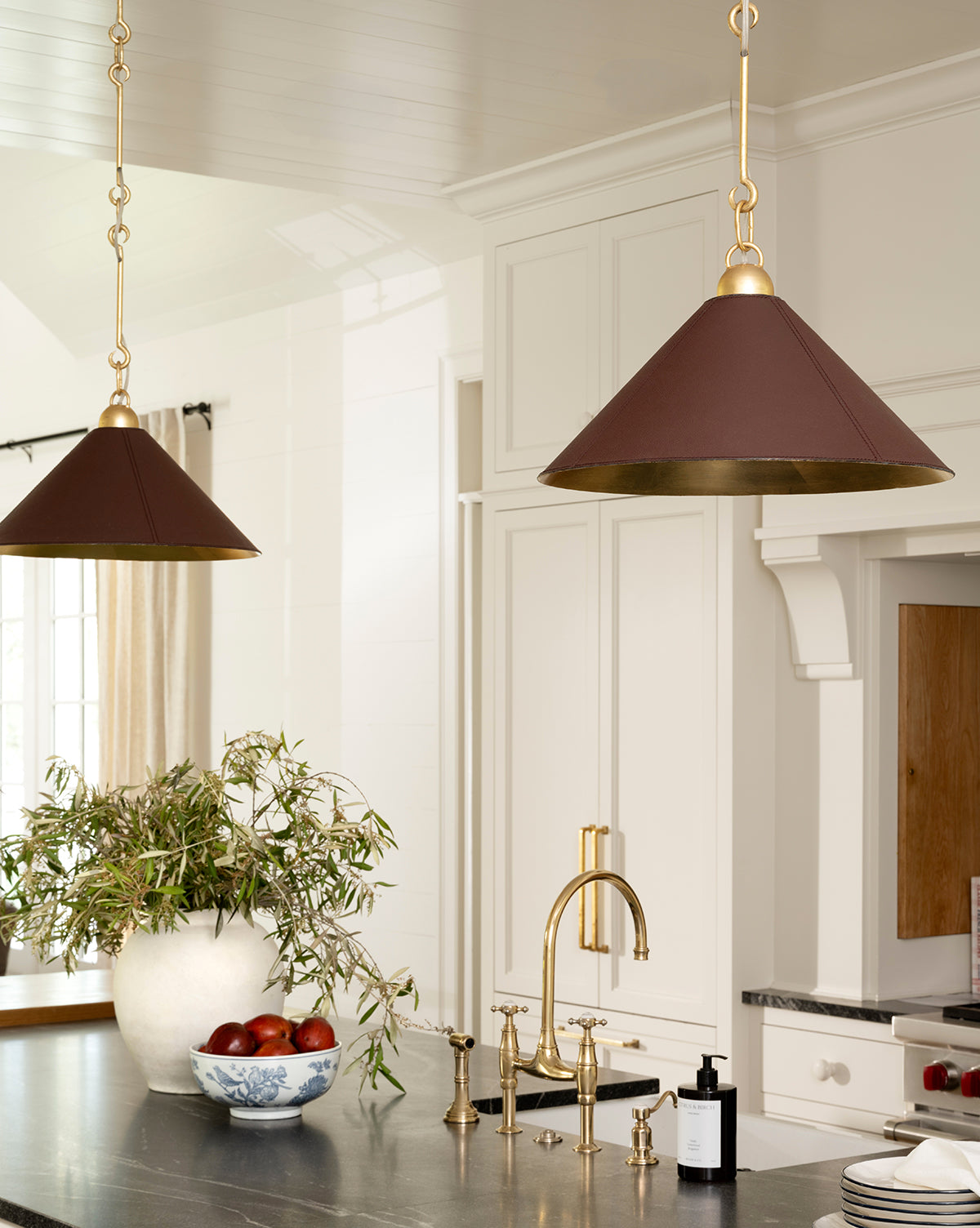 A bright kitchen features gold pendant lights, a black countertop, a gold faucet, and the Hand Stamped Botanical Bowl filled with apples atop white cabinetry beneath a window with airy curtains by McGee & Co.