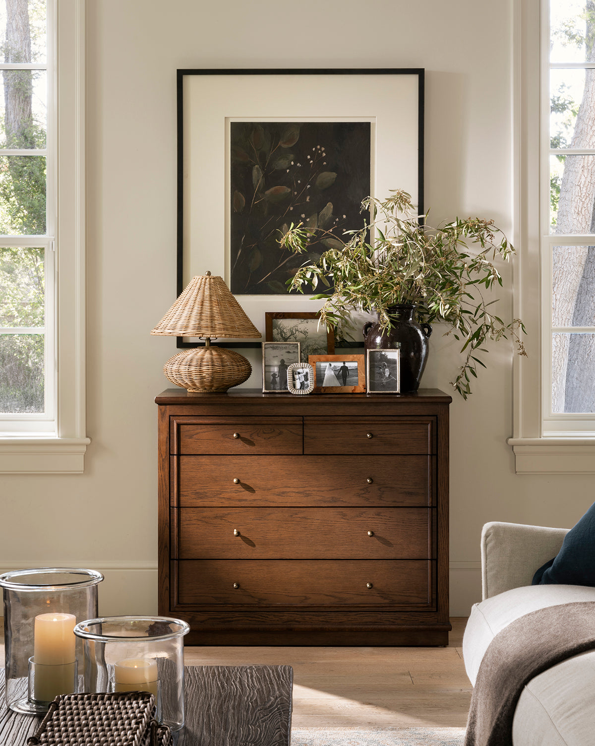 A Yorkshire Dresser with brass handles holds a lamp, framed photos, and a vase with greenery. Framed artwork sits behind it. Two large windows and a sofa add to this bright, stylish room—ideal for modern bedroom storage by McGee & Co.