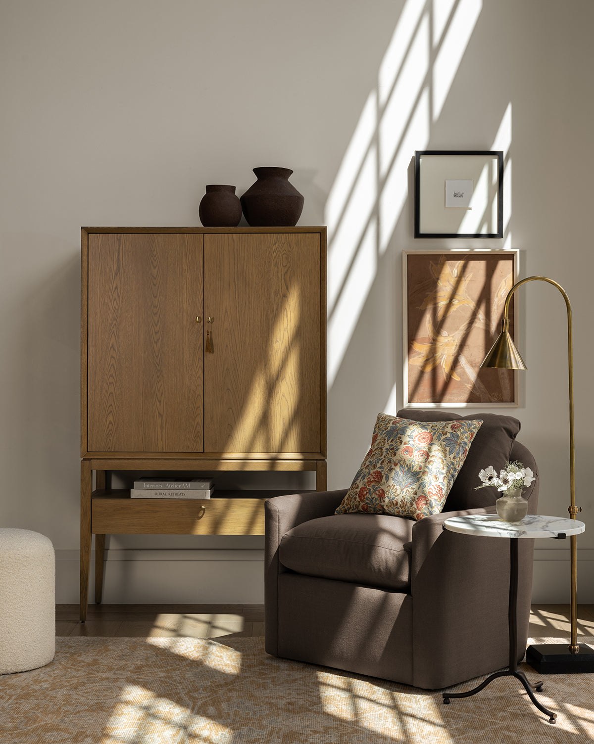 A cozy living space features a brown armchair with a floral pillow, the elegant Jahnsen Cabinet in honey oak, framed wall art, a round side table with flowers, a floor lamp, and sunlight streaming in through the window, by McGee & Co.
