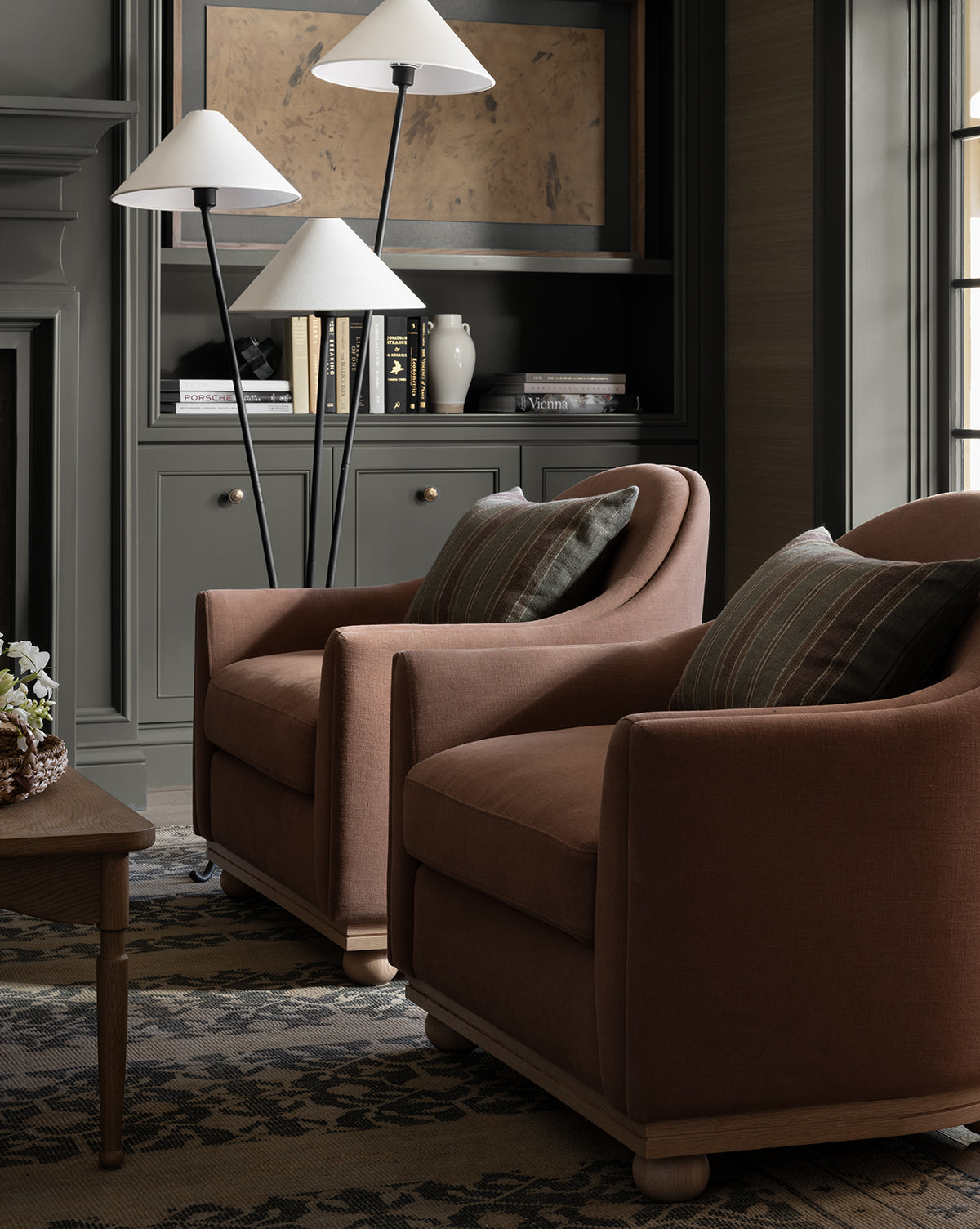Two Bunnell Lounge Chairs (Ready to Ship) with striped pillows are placed by a wooden coffee table in a cozy room. Behind them, a dark grey built-in bookshelf holds books, vases, and three white-shaded floor lamps, by McGee & Co.