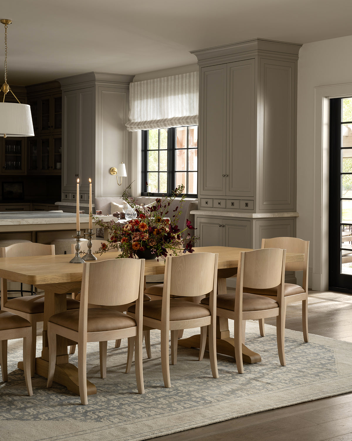 A modern dining room with a wooden table, eight Shepherd Dining Chairs with upholstered seats and white oak frames, a floral centerpiece, and candles. Gray cabinets, large windows with white curtains, and a patterned rug complete the look by McGee & Co.