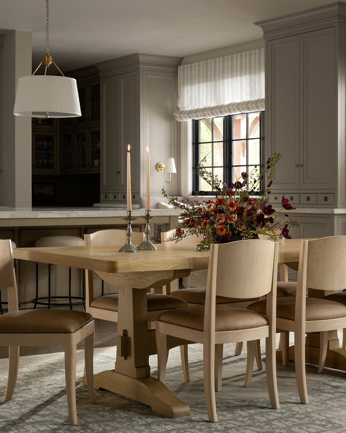 A cozy dining room features a Wilton Dining Table, six cushioned chairs, two glowing candlesticks, and a floral centerpiece. Soft light streams through a window with white blinds; neutral cabinets and a pendant light complete the scene, by McGee & Co.