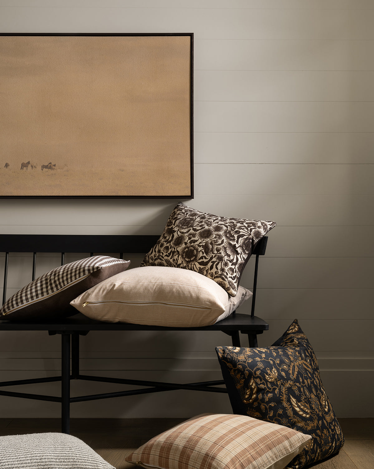 A black wooden bench holds patterned and plain throw pillows in neutral and dark tones. More pillows rest on the floor. Above the bench, Smokey Pasture beige minimalist giclee print hangs on a white shiplap wall, by McGee & Co.