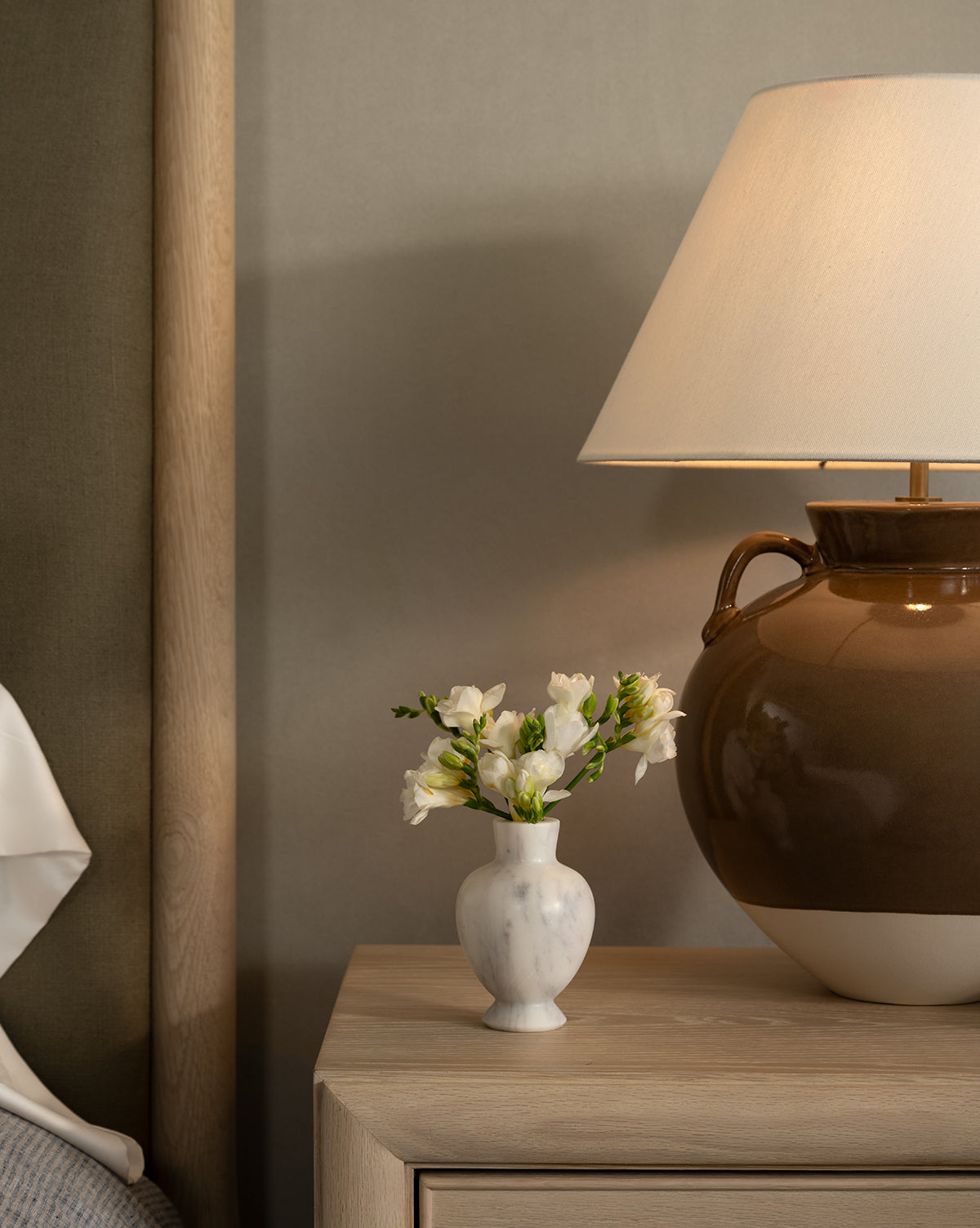 The Fera Bud Vase, made of white marble and filled with white flowers, sits on a light wooden nightstand beside a large brown and white ceramic lamp with a beige shade, set against a neutral wall near the bed, by McGee & Co.