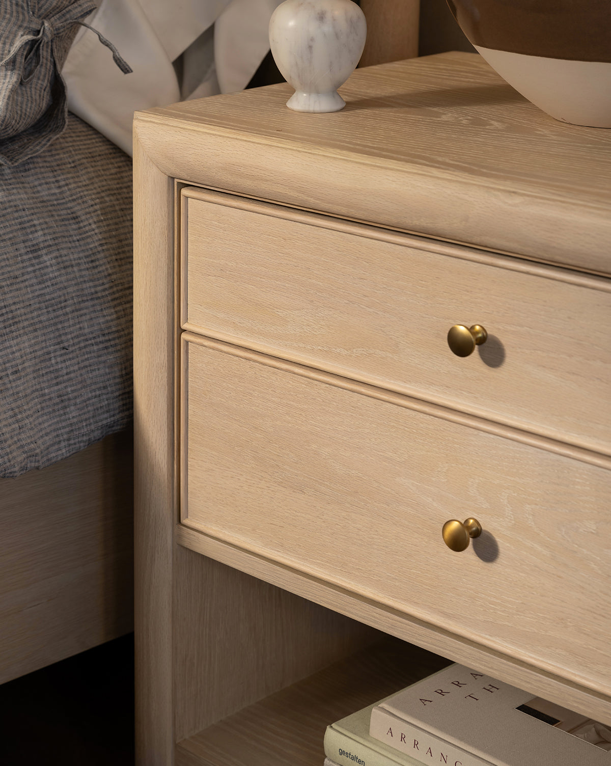 The Tellerbeck Nightstand in bleached oak, featuring two gold-knob drawers, brings elegance to your bedroom with a marble vase on top, books on the lower shelf, and a gray striped blanket draped over the nearby bed, by McGee & Co.