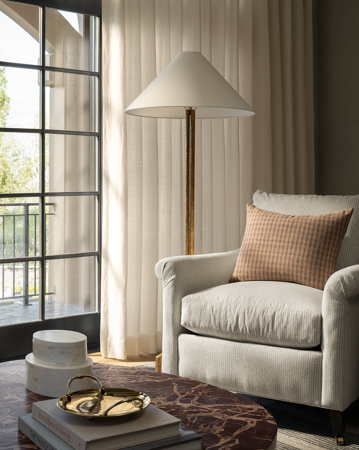 A cozy living room corner with a cream armchair, plaid cushion, Clifford Large Forged Floor Lamp with a white linen shade, marble coffee table, stacked books, decorative tray, and sheer curtains letting natural light in from big windows by McGee & Co.
