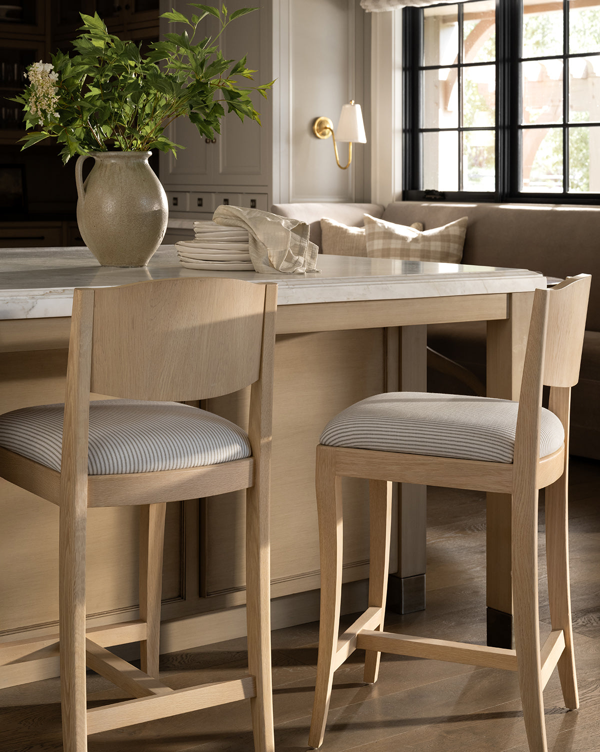 A sunlit kitchen features a marble-topped island with two Makerspalm Shepherd Counter Stools, a ceramic pitcher with greenery, stacked plates, and a built-in bench by large windows—McGee & Co.