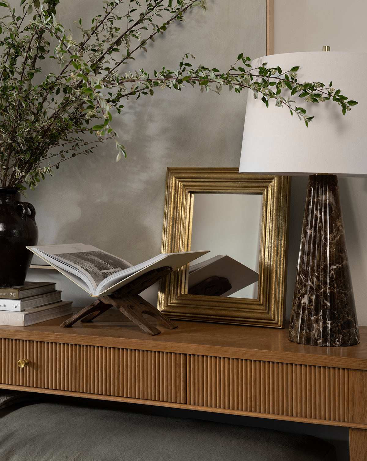A wooden console table displays an open book on a stand, stacked books, a black vase with leafy branches, the Suzanne Shelf Mirror in a gold-finished frame, and a marble-based lamp with a white shade by McGee & Co.