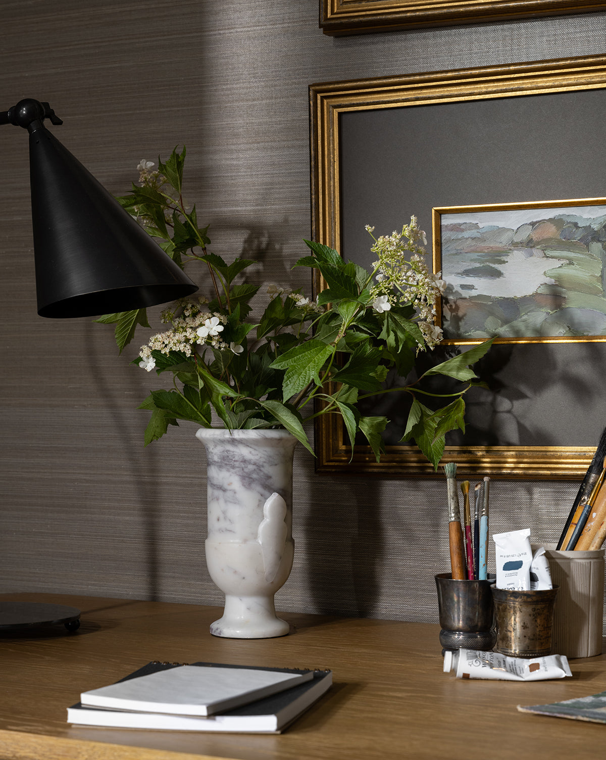 The Rayla Marble Vase, filled with white flowers and greenery, sits on a wooden desk beside a black lamp, art supplies, a closed notebook, and framed art—ideal for adding elegant traditional decor to any space, by McGee & Co.