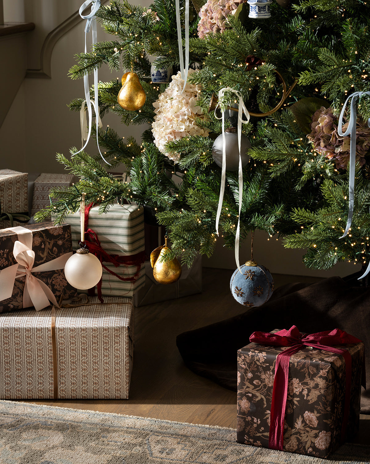 The Chateau Noel Tree, decorated with white and gold ornaments, ribbons, and fairy lights, stands above a velvet tree skirt with several wrapped presents and bows arranged nearby, by McGee & Co.