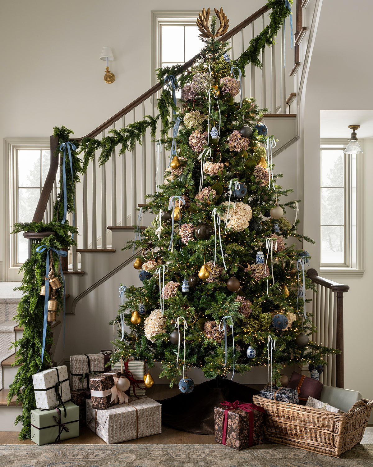 A Chateau Noel Tree adorned with ribbon and ornaments stands by a staircase draped in garland, its velvet skirt at the base with wrapped presents, a yarn basket, and sunlight streaming through nearby windows, by McGee & Co.