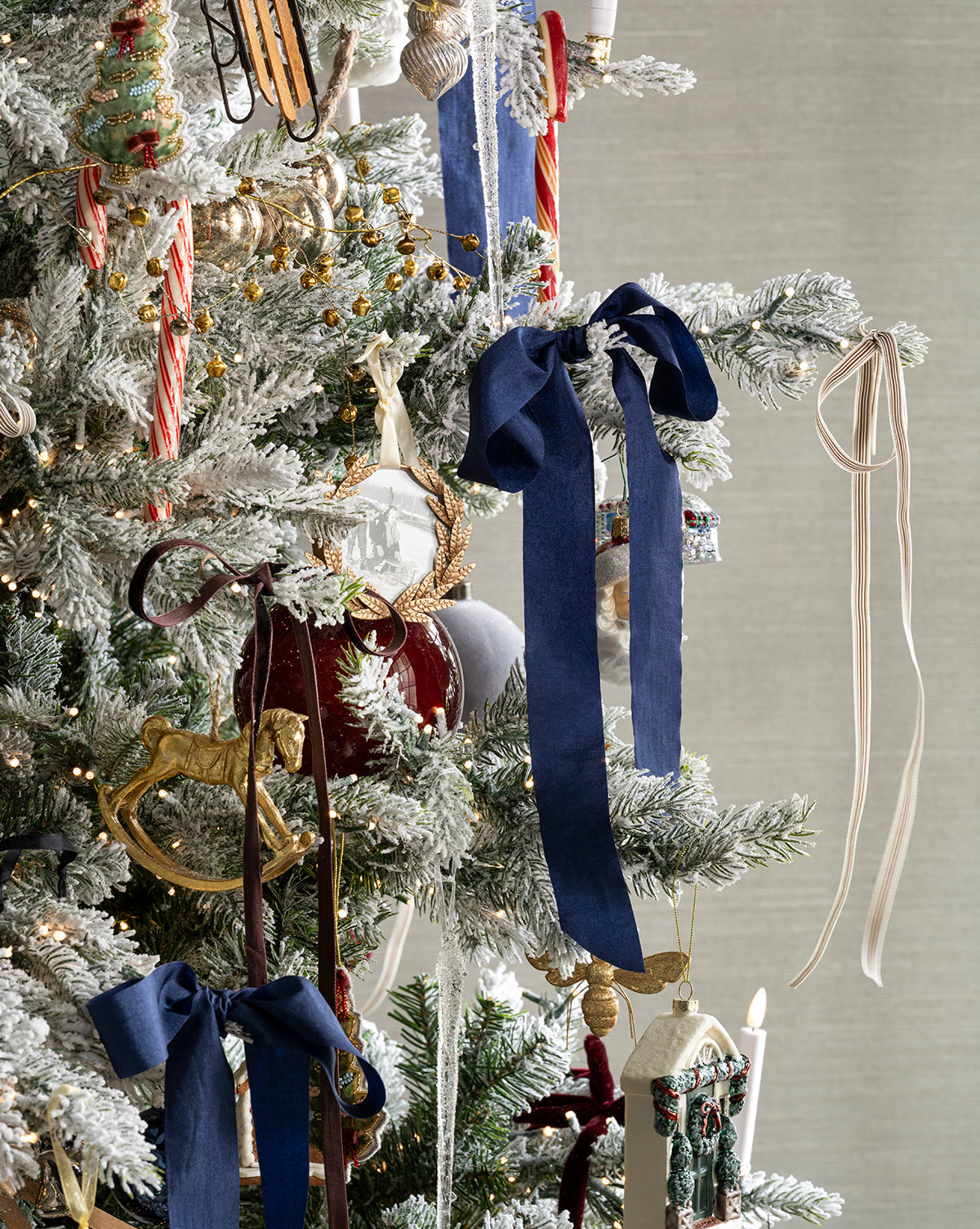 The Heritage Holiday Tree, snow-frosted and adorned with blue bows, candy canes, gold ornaments, picture frames, rocking horse, striped ribbons, and hanging candles set against a neutral background, by McGee & Co.
