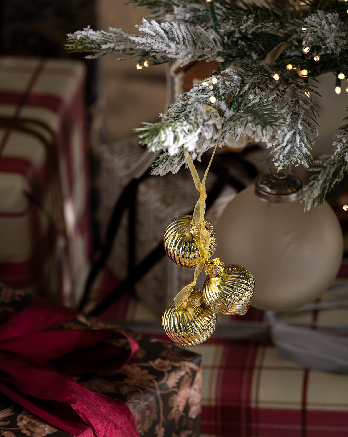 A close-up of a snow-dusted Christmas tree branch features three Glass Bundle Ornaments (Set of 4) hanging from yellow ribbons, surrounded by gifts in plaid and floral paper and warm fairy lights, by McGee & Co.