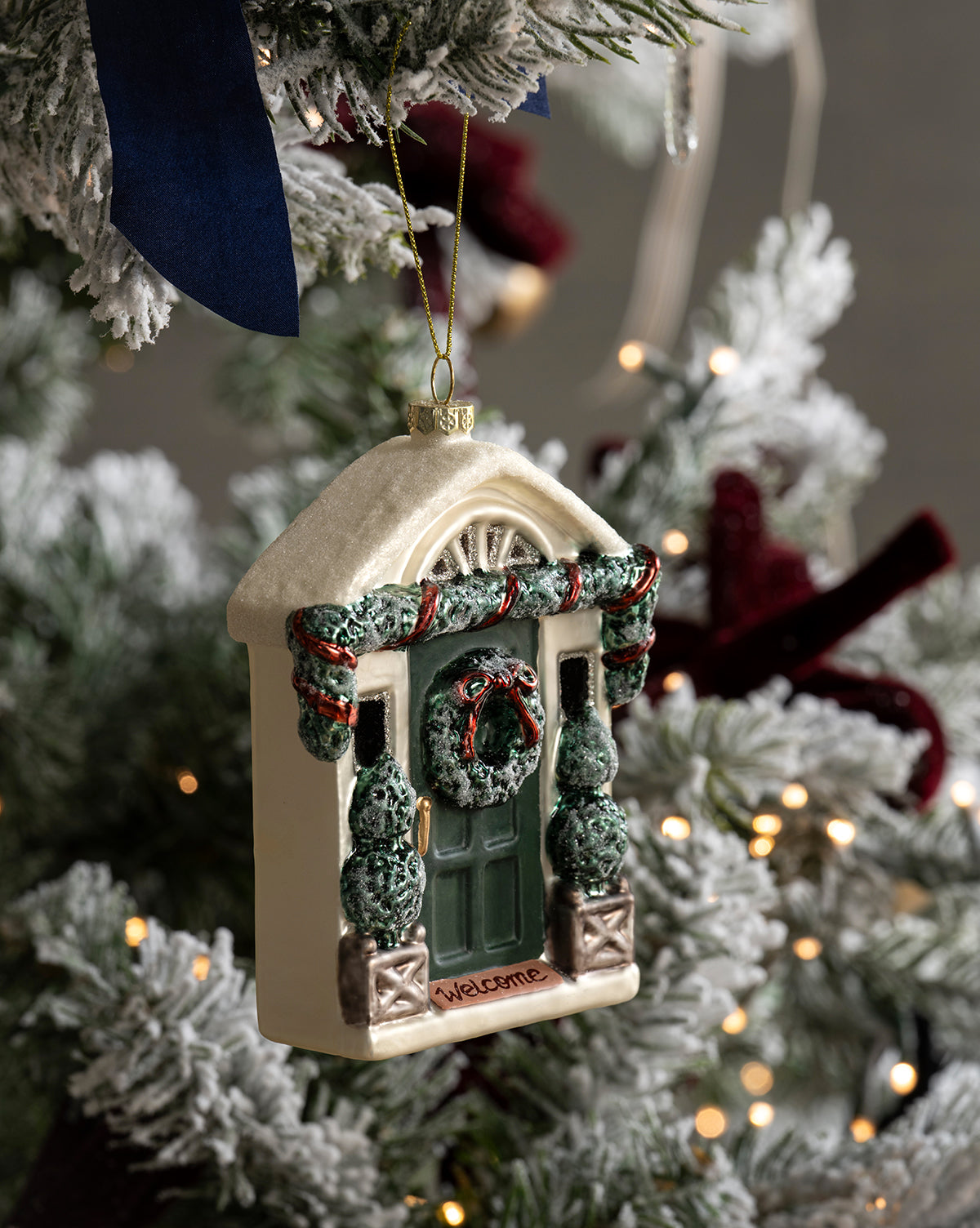 A house-shaped ornament with a green door, wreath, and garland hangs on the snow-frosted Heritage Holiday Tree, adorned with twinkling lights, glass baubles, and a dark blue ribbon, by McGee & Co.