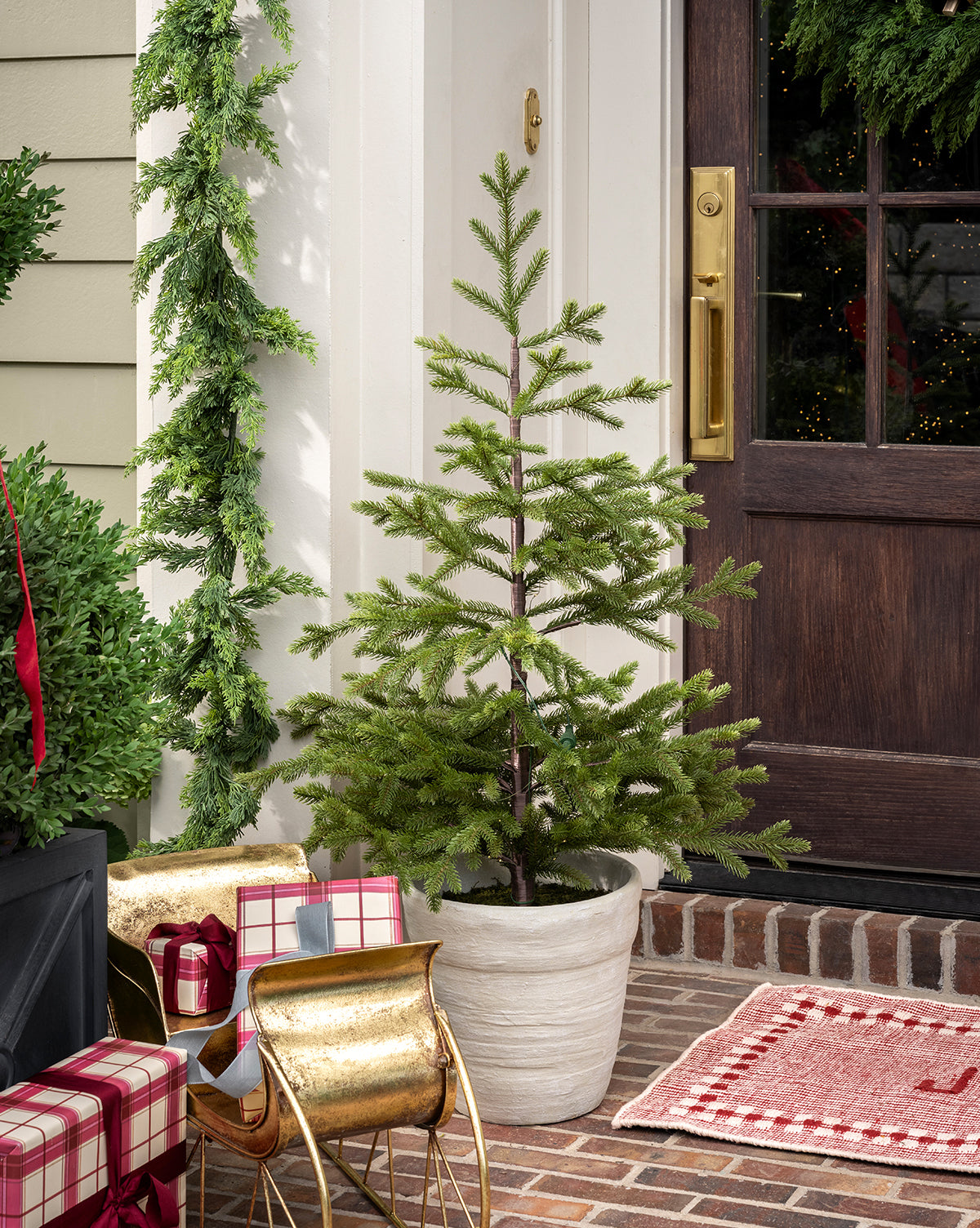 The Faux Spruce Pre-Lit Potted Tree stands on a brick porch by a dark wooden door, accented with wrapped gifts, a golden sleigh, festive garland, and a red plaid rug for a cozy holiday ambiance, by McGee & Co.