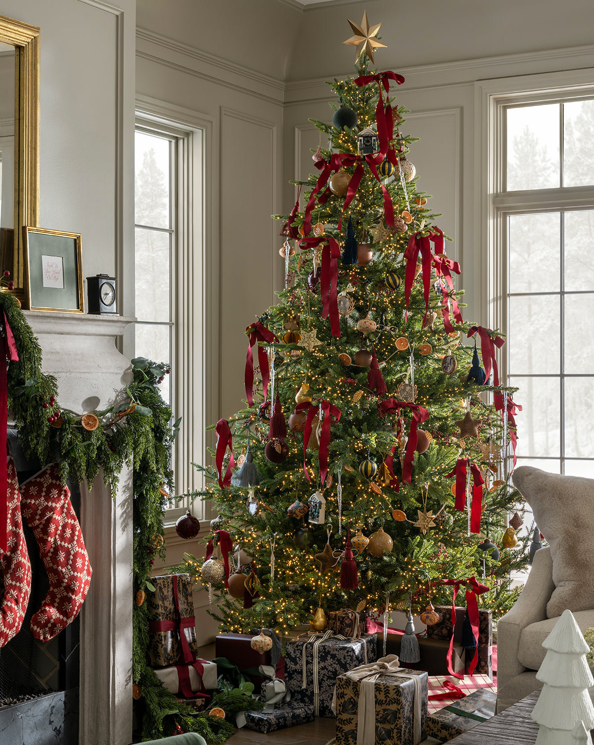 The Classic Christmas Tree, inspired by the Grandis Fir, stands by the window decorated with red ribbons, ornaments, and a gold star. Below rest presents; nearby, stockings and garland adorn the mantel in a sunlit living room, by McGee & Co.