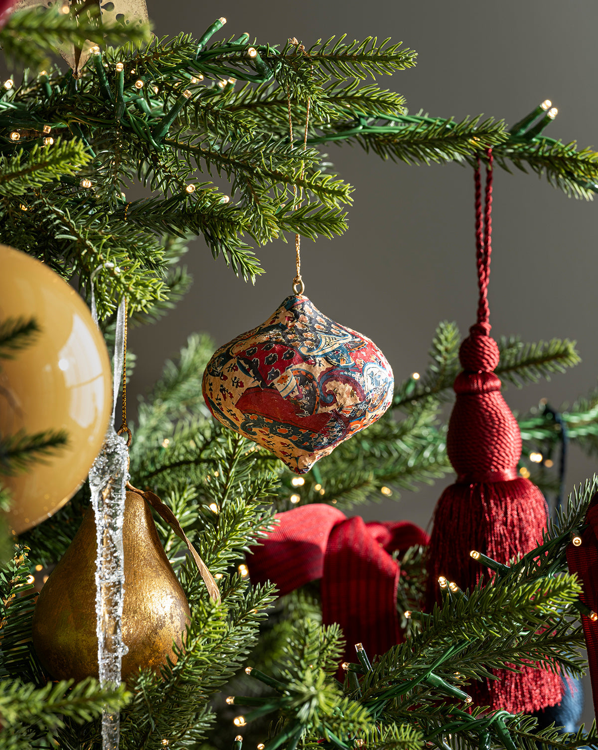 Close-up of the Classic Christmas Tree highlighting a designer mix of colorful patterned ornaments, a gold pear, clear icicle, red tassels, and warm white lights on lush branches, by McGee & Co.