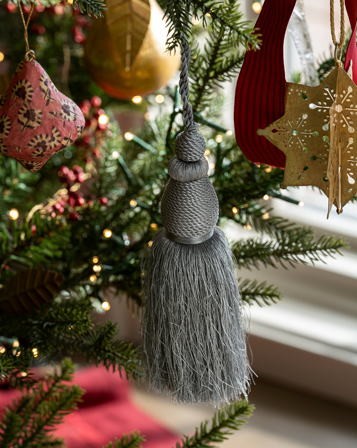 The Lucia Bullion Tassel hangs on a festive Christmas tree with green branches, twinkling lights, and ornaments like a gold star and red fabric shape, bringing vintage-inspired charm to your holiday décor, by McGee & Co.