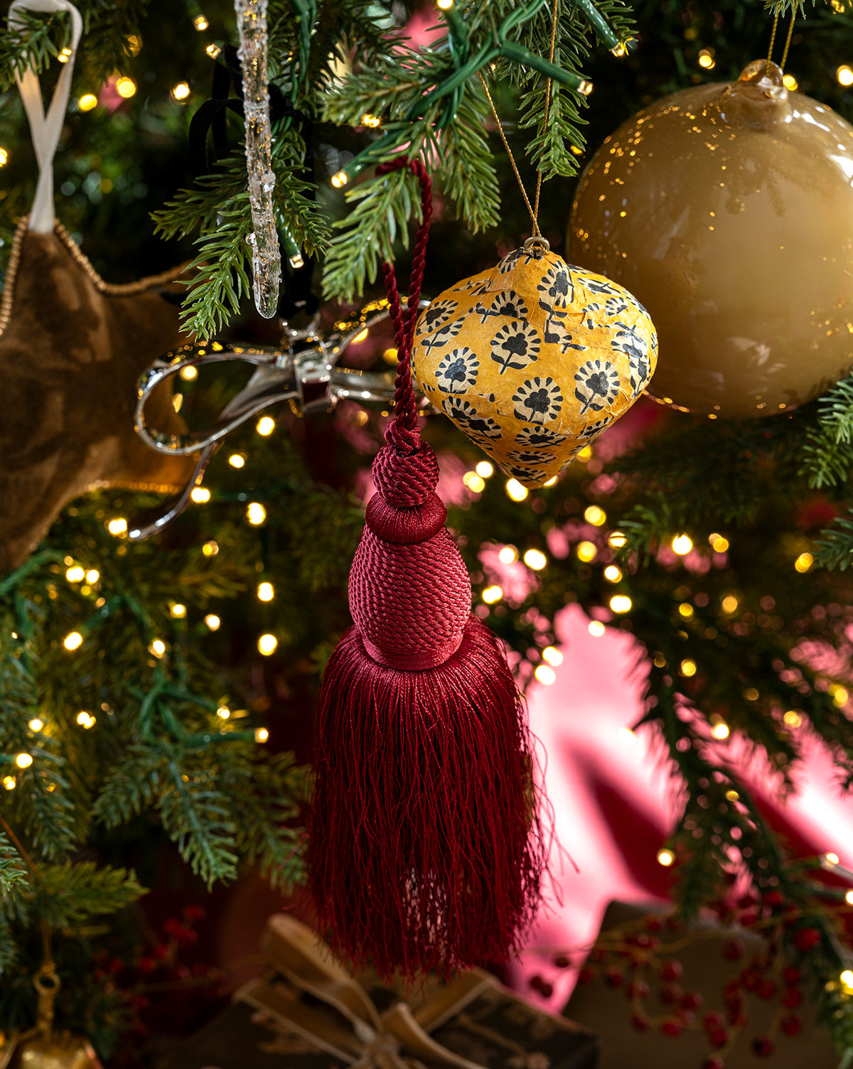 A close-up of a decorated Christmas tree featuring the maroon Lucia Bullion Tassel ornament, a yellow patterned bauble, gold ornaments, twinkling fairy lights, green pine branches, and a wrapped gift at the base, by McGee & Co.