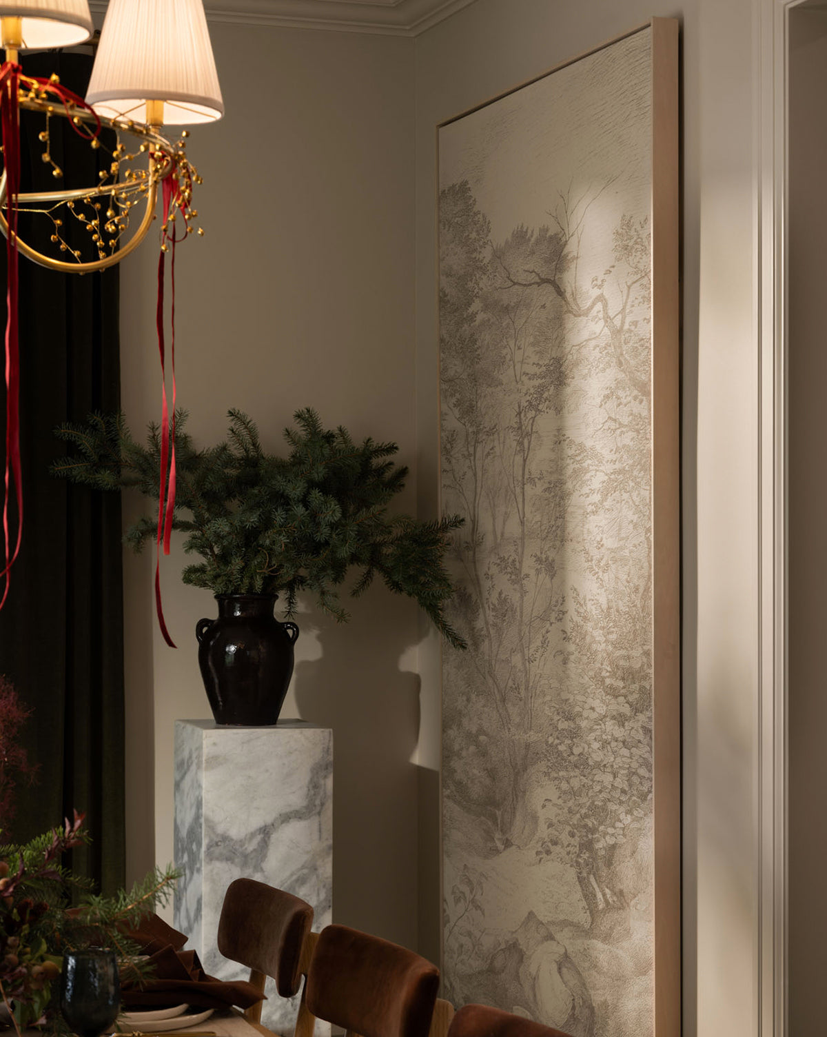 A dimly lit dining room corner features a black vase with greenery on a marble pedestal, beside the Neutral Toile No. 2 gallery-quality canvas art of a landscape sketch, with a light fixture adorned with red ribbons overhead, by McGee & Co.