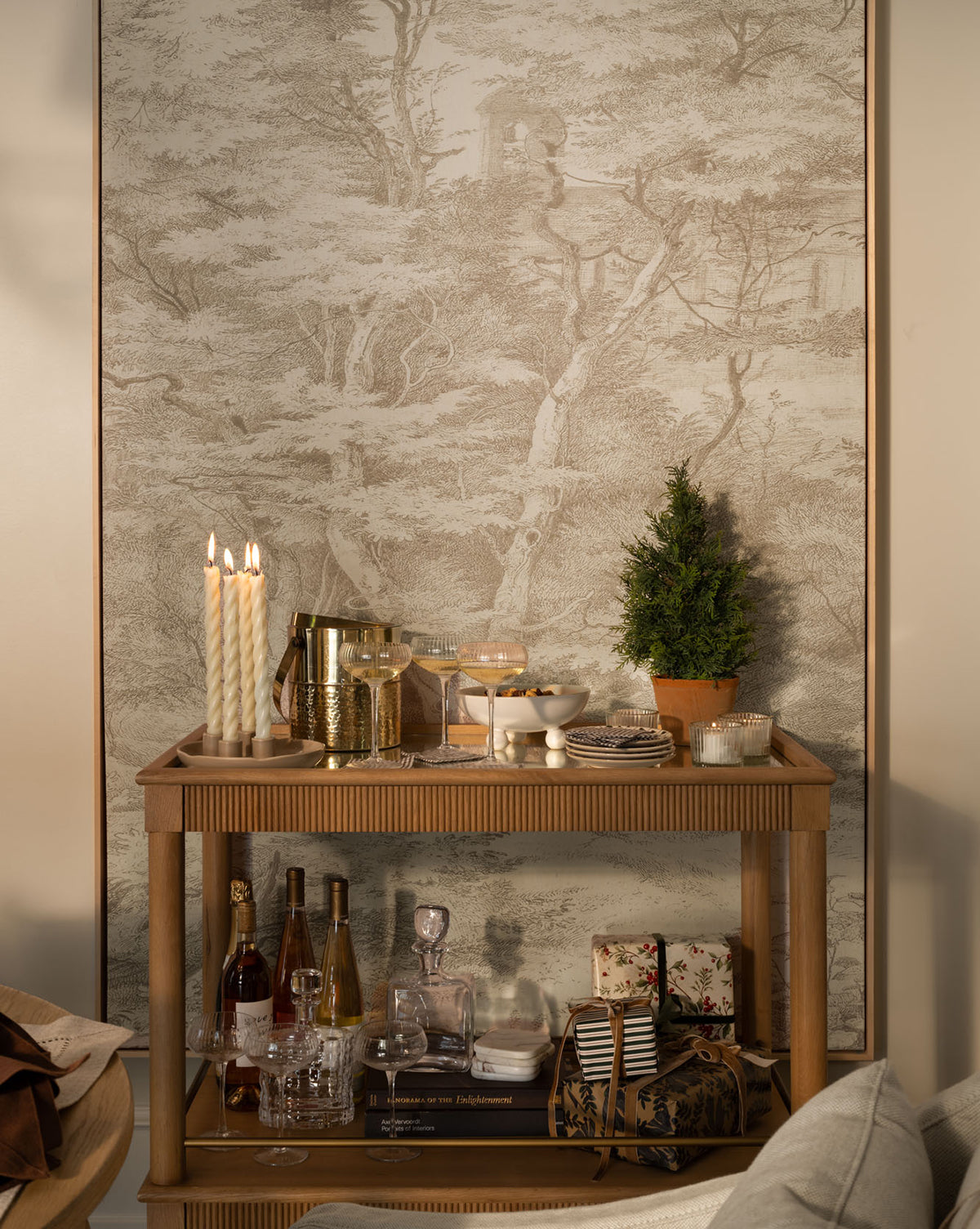 A wooden bar cart holds drinks, glasses, gifts, a plant, and candles, set against the Neutral Toile No. 1 gallery-quality giclee print mural featuring trees and a building in the background, by McGee & Co.