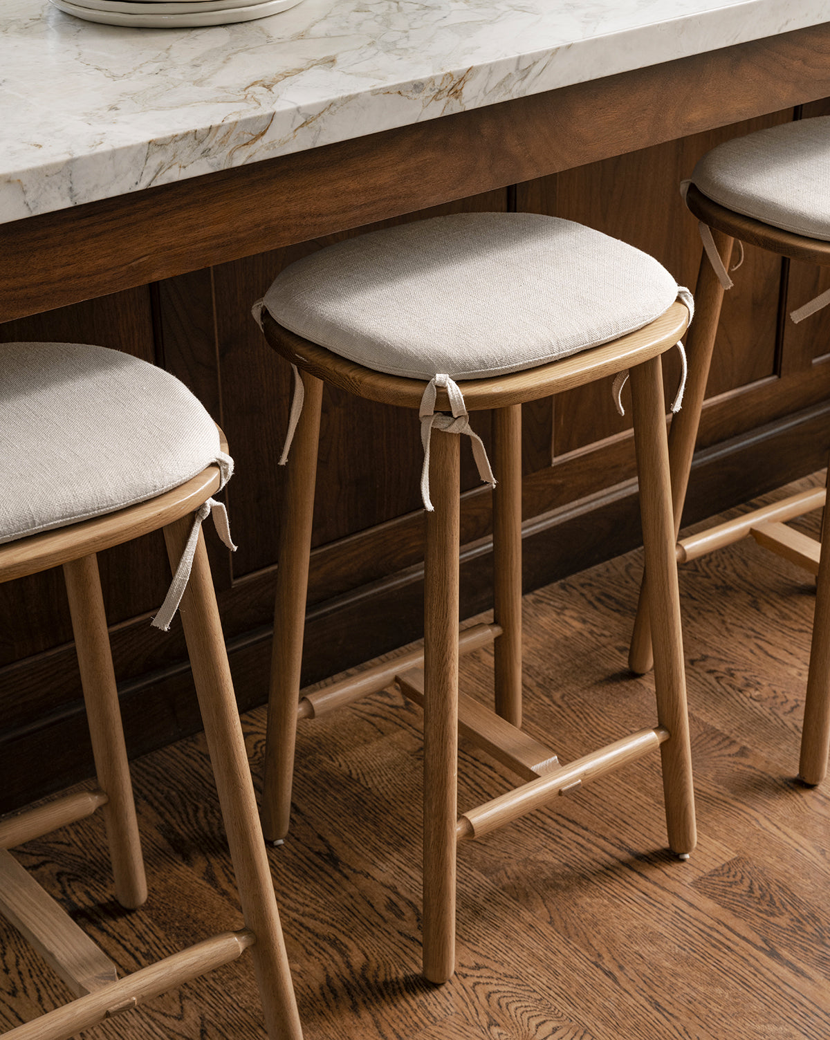 Three Ralph Oak Counter Stools with linen cushions and ribbon ties sit under a marble kitchen counter with dark wood paneling, all on a wooden floor by McGee & Co.