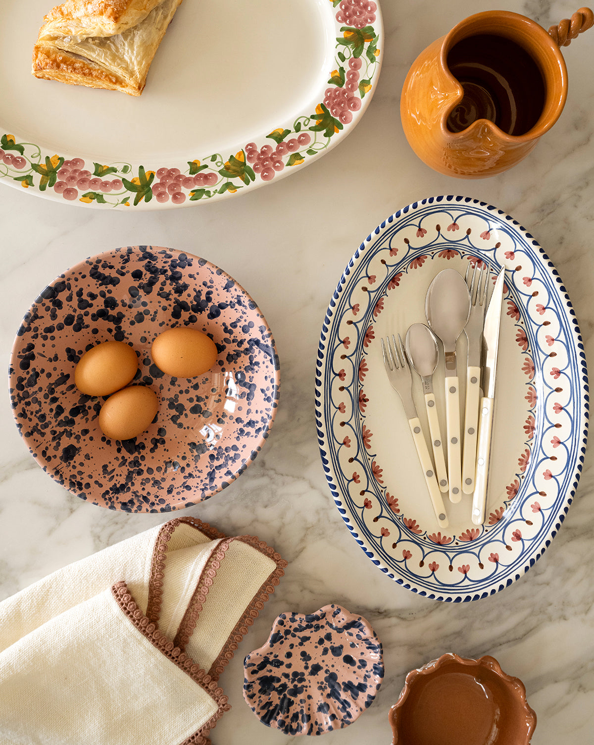 A marble table displays a slice of pastry on a floral plate, three brown eggs in the Splatter Large Bowl, a patterned oval dish with cutlery, a brown jug, napkins, and small matching dishes by McGee & Co.