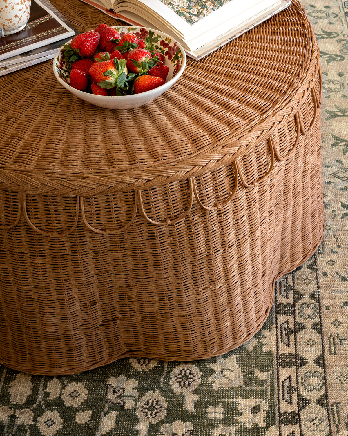 A round wicker table with a scalloped edge holds the Vine Large Bowl filled with strawberries and an open book, set on a green and beige patterned rug, by McGee & Co.