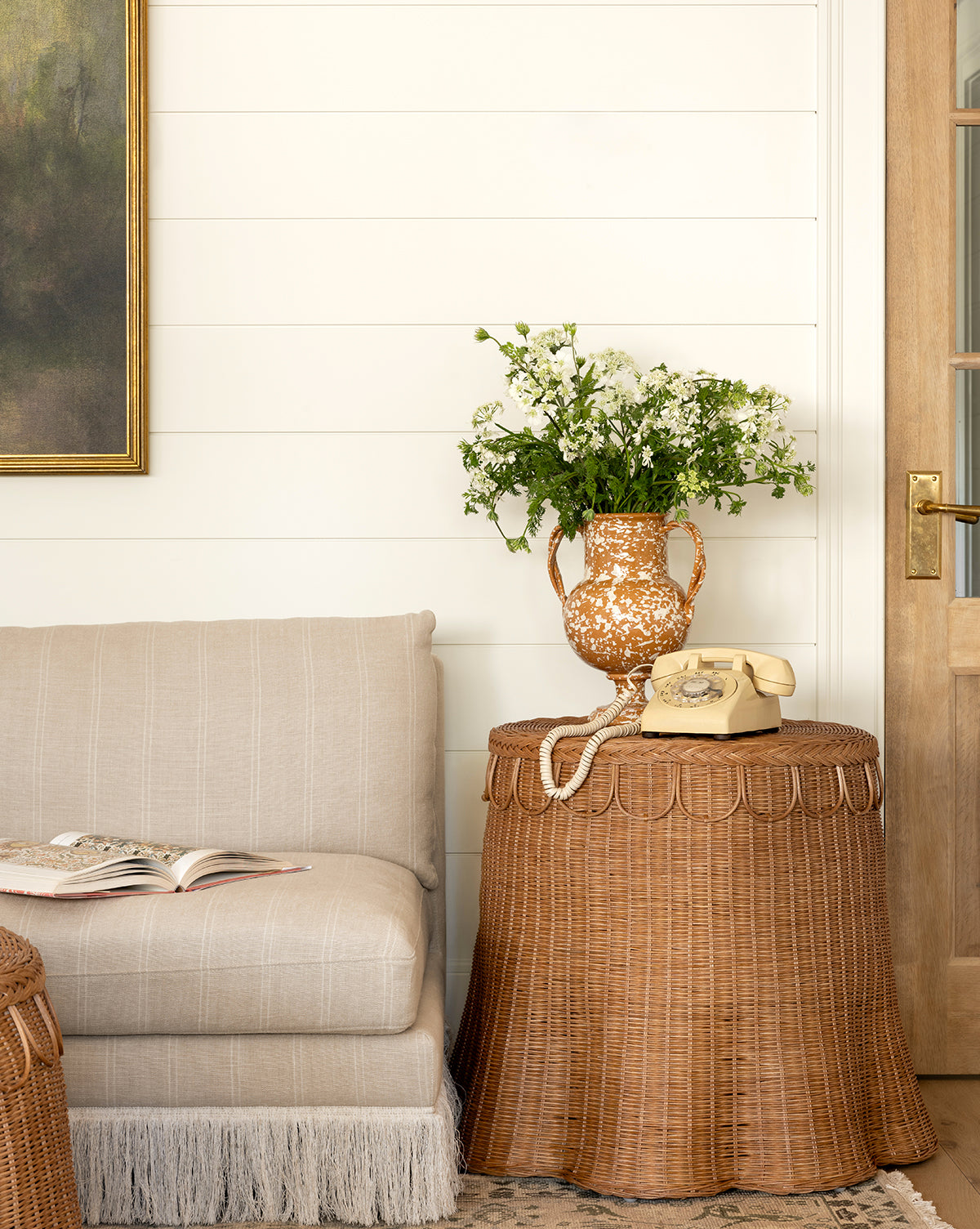 A cozy living room corner with a beige loveseat, an open book, and the Adeline Side Table by Sharland England LTD topped with a yellow rotary phone and white flowers, beside a wooden door and a painting on a white shiplap wall, McGee & Co.