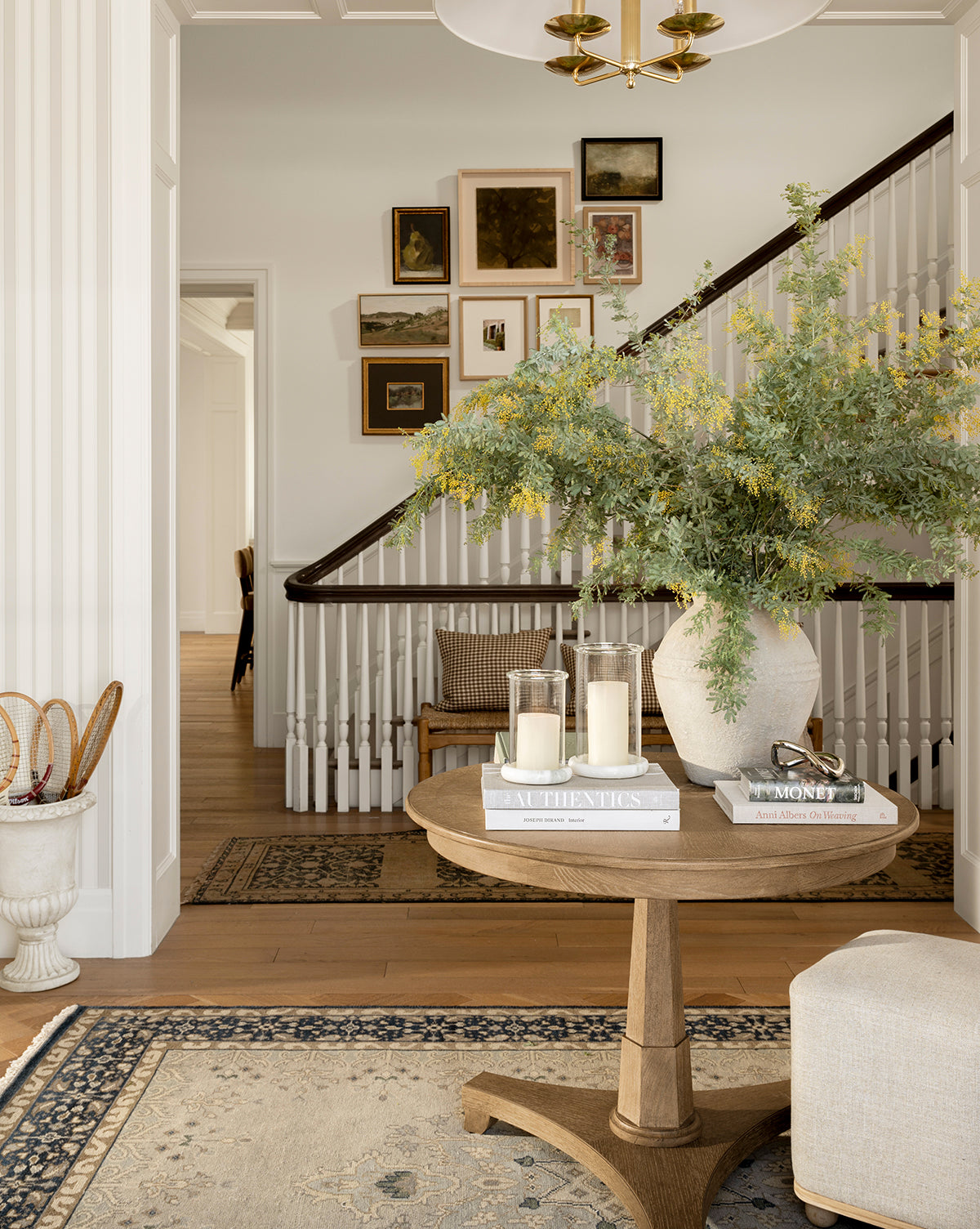 A sunlit entryway includes the Marna Entry Table with a round top, styled with a vase of greenery, books, and candles. Nearby, a staircase showcases framed art and a patterned rug for an inviting, elegant look by McGee & Co.