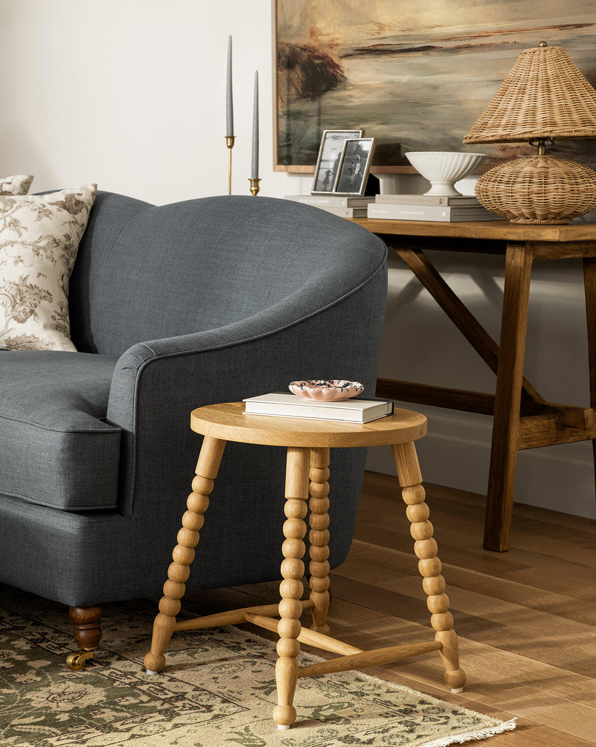 A cozy living room with a dark gray sofa, the Claire Side Table featuring beaded legs holding a book and small dish, and a wooden console table with a lamp, photos, and decor accents in the background by McGee & Co.