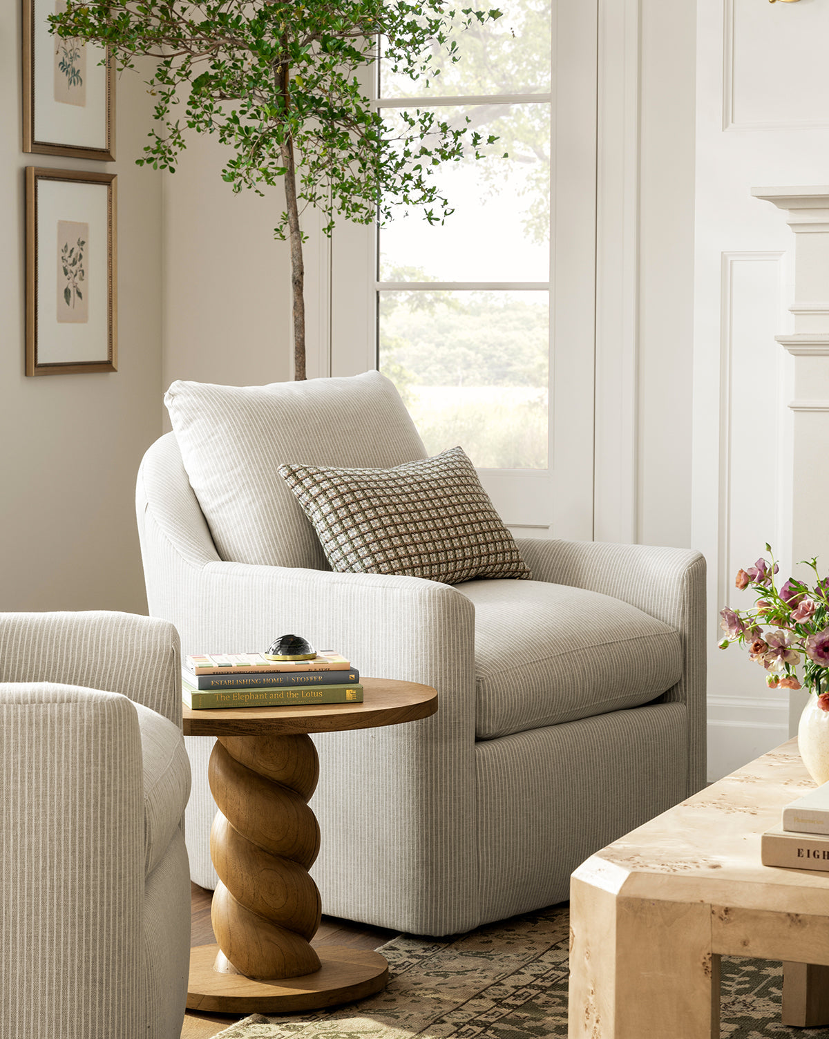 A cozy living room with a light gray striped armchair, a patterned pillow, a round wooden side table with books, a potted plant, and Delicate Floral lll art in a silver wood frame gracing the wall by the window’s natural light.