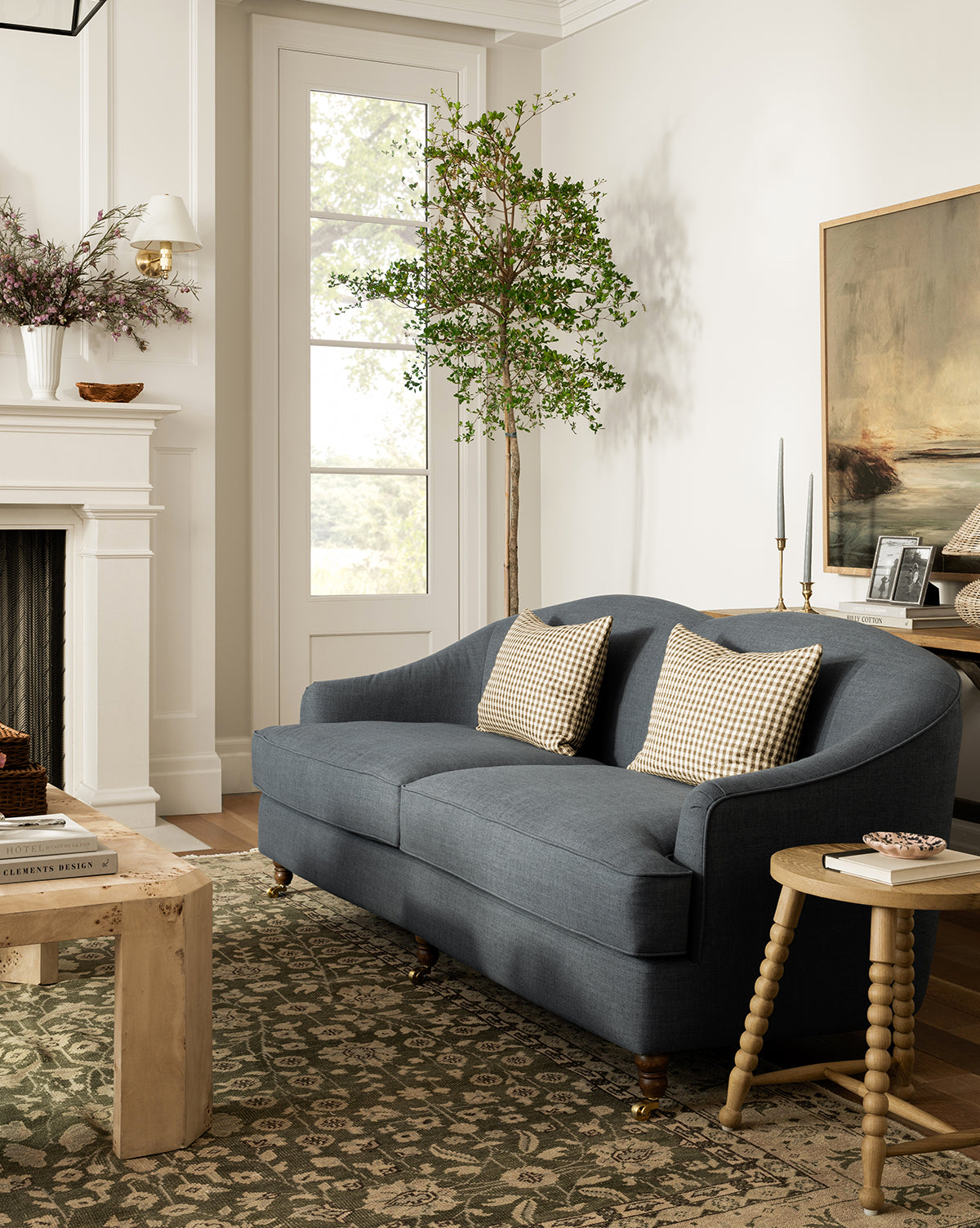 A cozy living room featuring a blue Fletcher Sofa 76 with two patterned pillows, a round wooden side table, green patterned rug, potted tree, fireplace, wall art, and abundant natural light filling the space by McGee & Co.