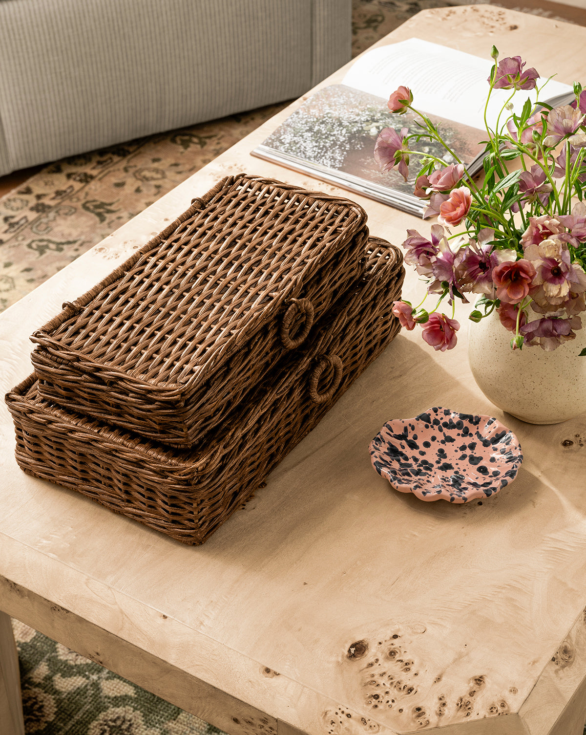 Two stacked Windmere Woven Boxes, a ceramic vase with pink and purple flowers, a speckled pink dish, and an open book rest on a light wooden coffee table in a cozy living room, by McGee & Co.
