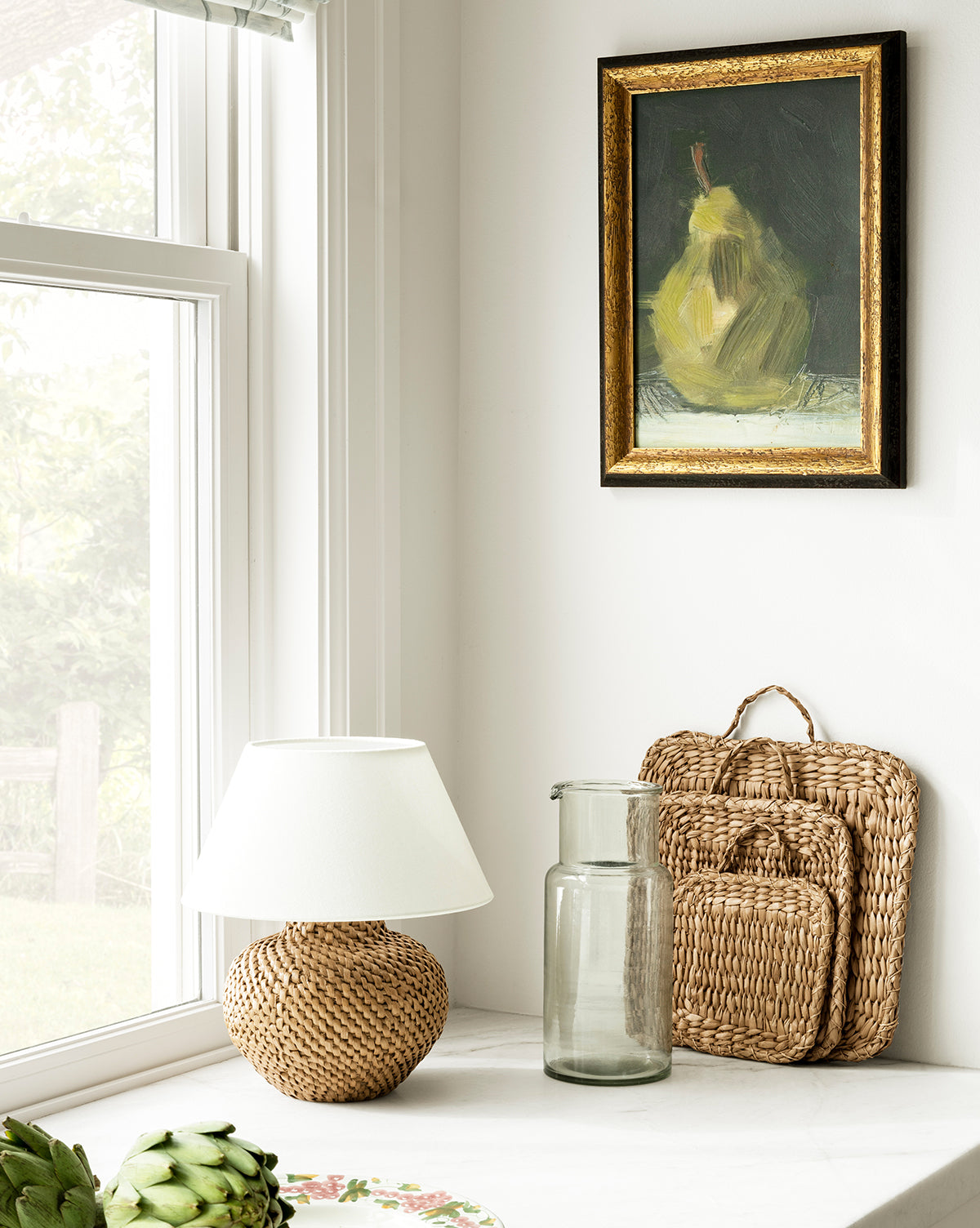A bright windowsill features a wicker lamp, woven trays, and artichokes. The Sienna Carafe sits beside a framed pear painting, creating an elegant dining display against the white wall, by McGee & Co.