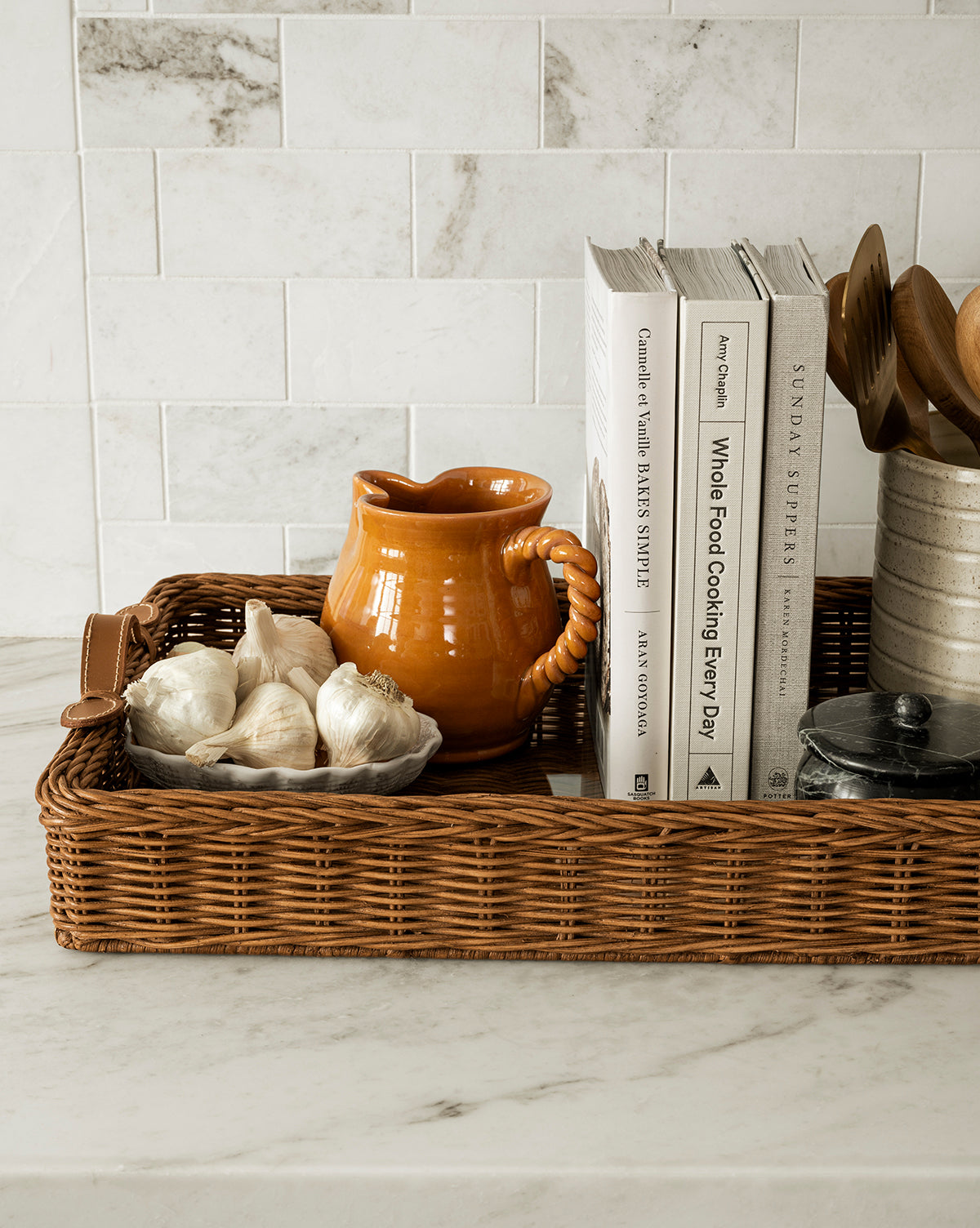 A wicker tray on a marble countertop holds garlic bulbs, the orange earthenware Louise Jug with a twisted handle, cookbooks, wooden utensils in a container, and a small black dish. A white tile backsplash appears in the background, by McGee & Co.