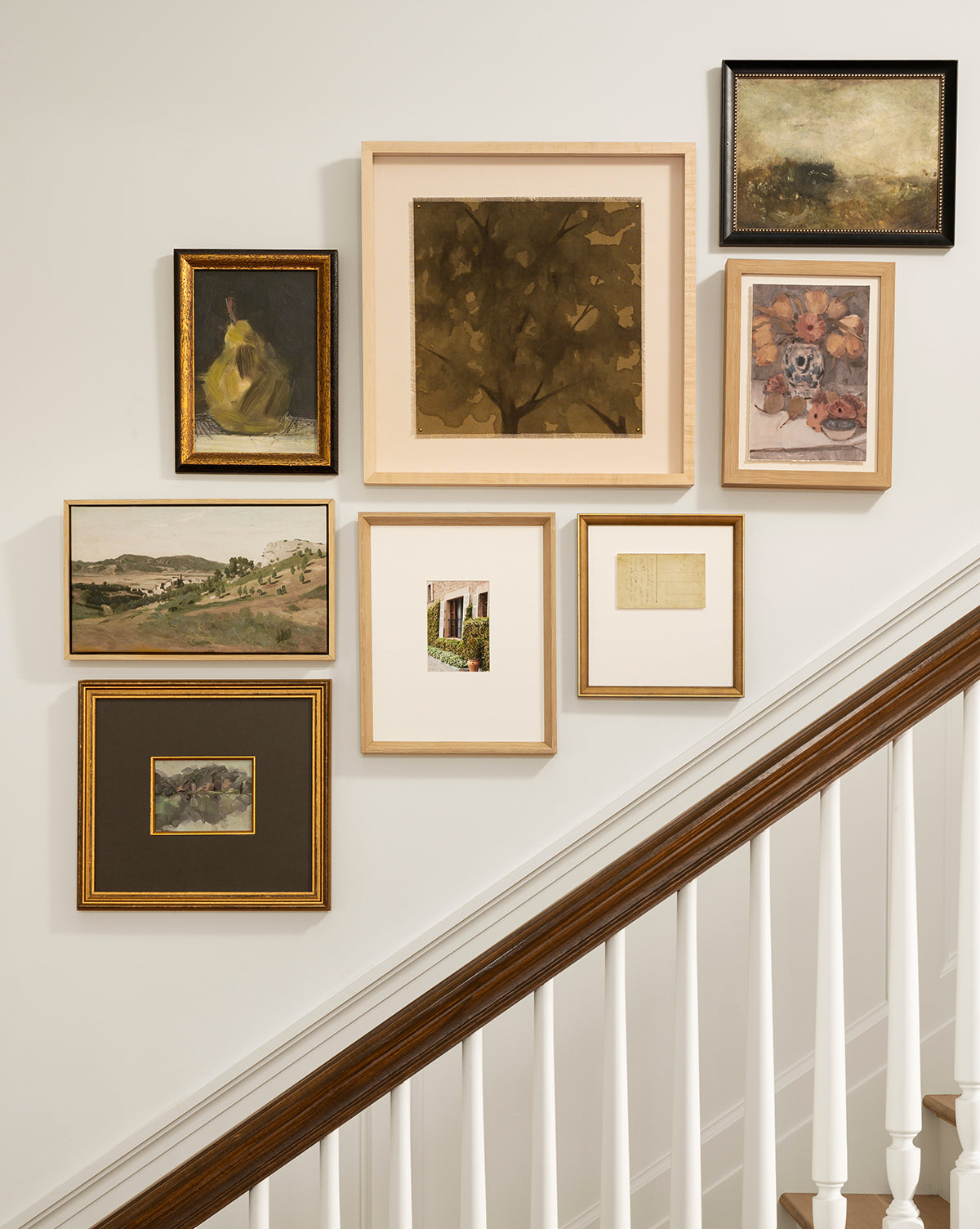 A gallery wall of seven framed artworks, including the Pear Pictorial and giclee prints, featuring landscapes and abstracts in neutral earthy tones, arranged above a wooden staircase with white railings, by McGee & Co.