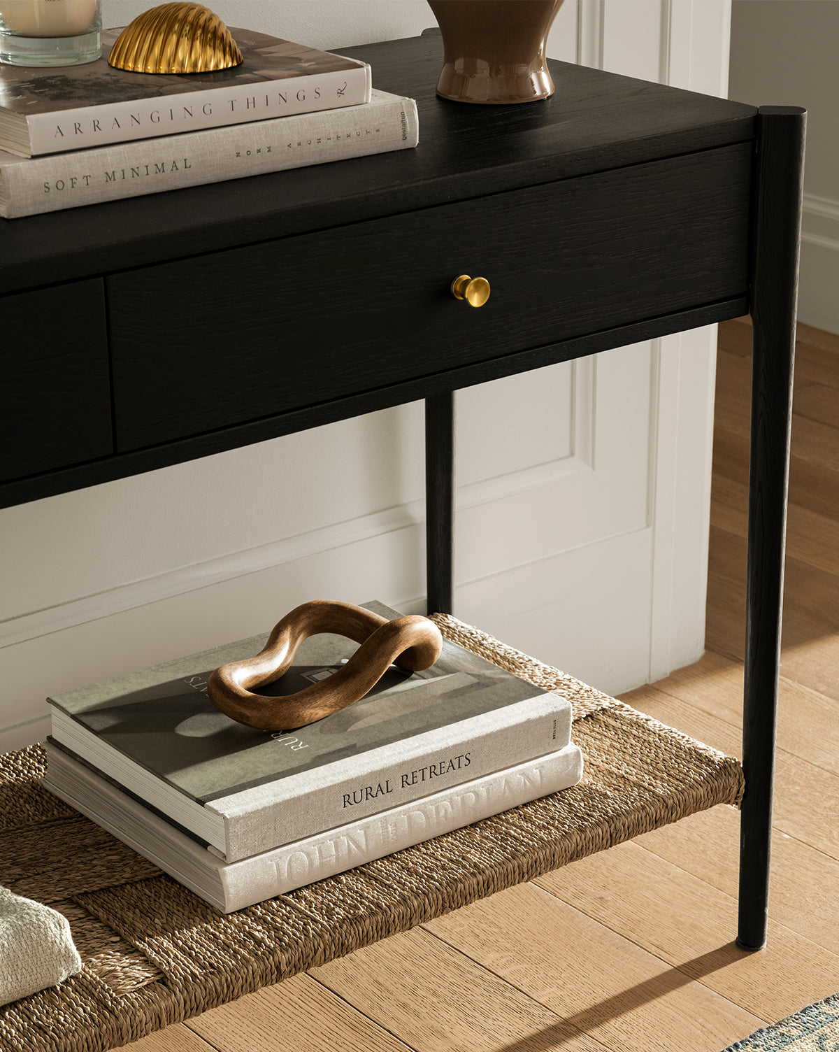 The Dana 2-Drawer Console in black with brass knobs stands on a wooden floor. Books and decor sit on top and a woven lower shelf, making it ideal for stylish entryways. Featured: Soft Minimal and Rural Retreats, by McGee & Co.