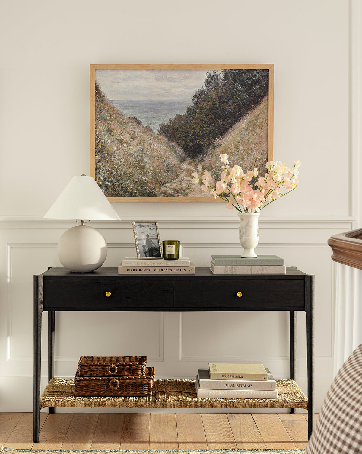 A black console table holds a white lamp, framed photo, books, and a vase of pink flowers. Above, Carved Path—displayed in a natural oak frame—hangs on the white wall; baskets and books are arranged on the floor below, by McGee & Co.
