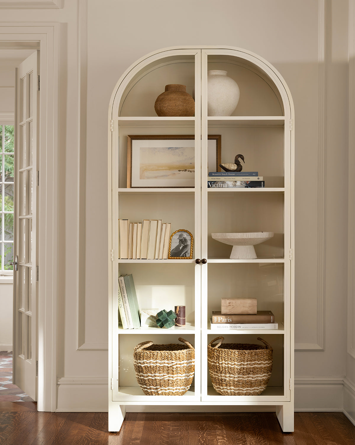 The white glass door Brookes Cabinet holds books, vases, framed art, a ceramic bird, a white pedestal bowl, and two woven baskets on the bottom shelf. Sunlight from a window highlights its timeless design by McGee & Co.