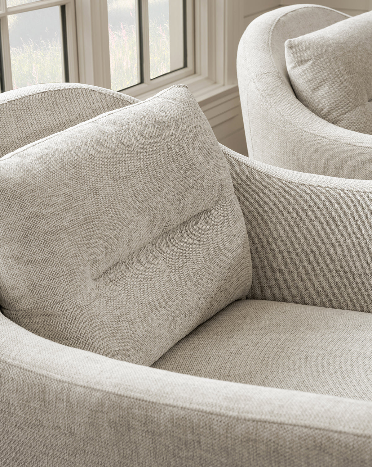 A close-up of the light beige Commodore Lounge Chair with twine crypton upholstery, a curved backrest, and cushion sits by a sunlit window. A second matching, deep-seated Commodore Lounge Chair is partially visible in the background, by McGee & Co.