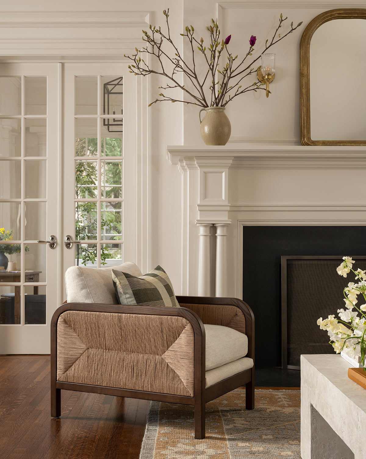 A Garvey Lounge Chair with woven sides and upholstered cushions sits by a white fireplace, while a vase on the mantel and French doors add light to this elegant, neutral-toned room, by McGee & Co.