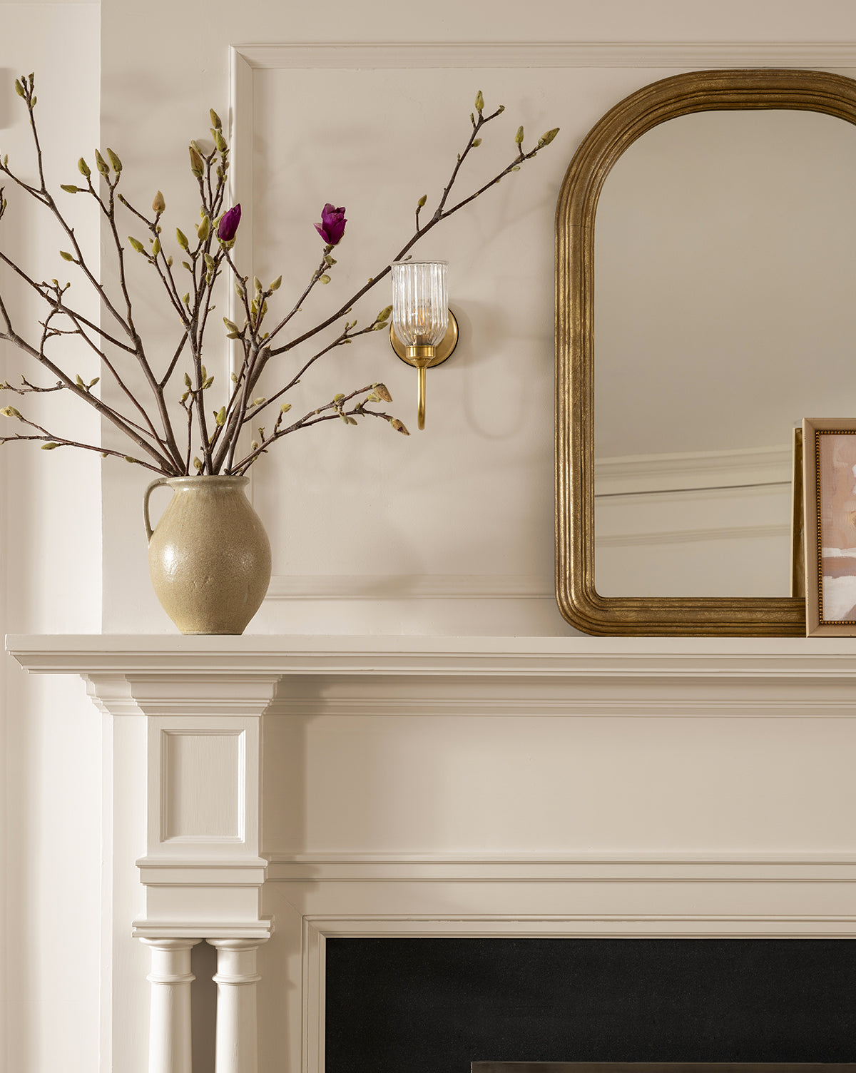 A beige fireplace mantel with a Raiden Vase filled with pink budding branches, a gold-framed mirror, wall sconce, and framed picture brings rustic charm to the neutral-toned room.