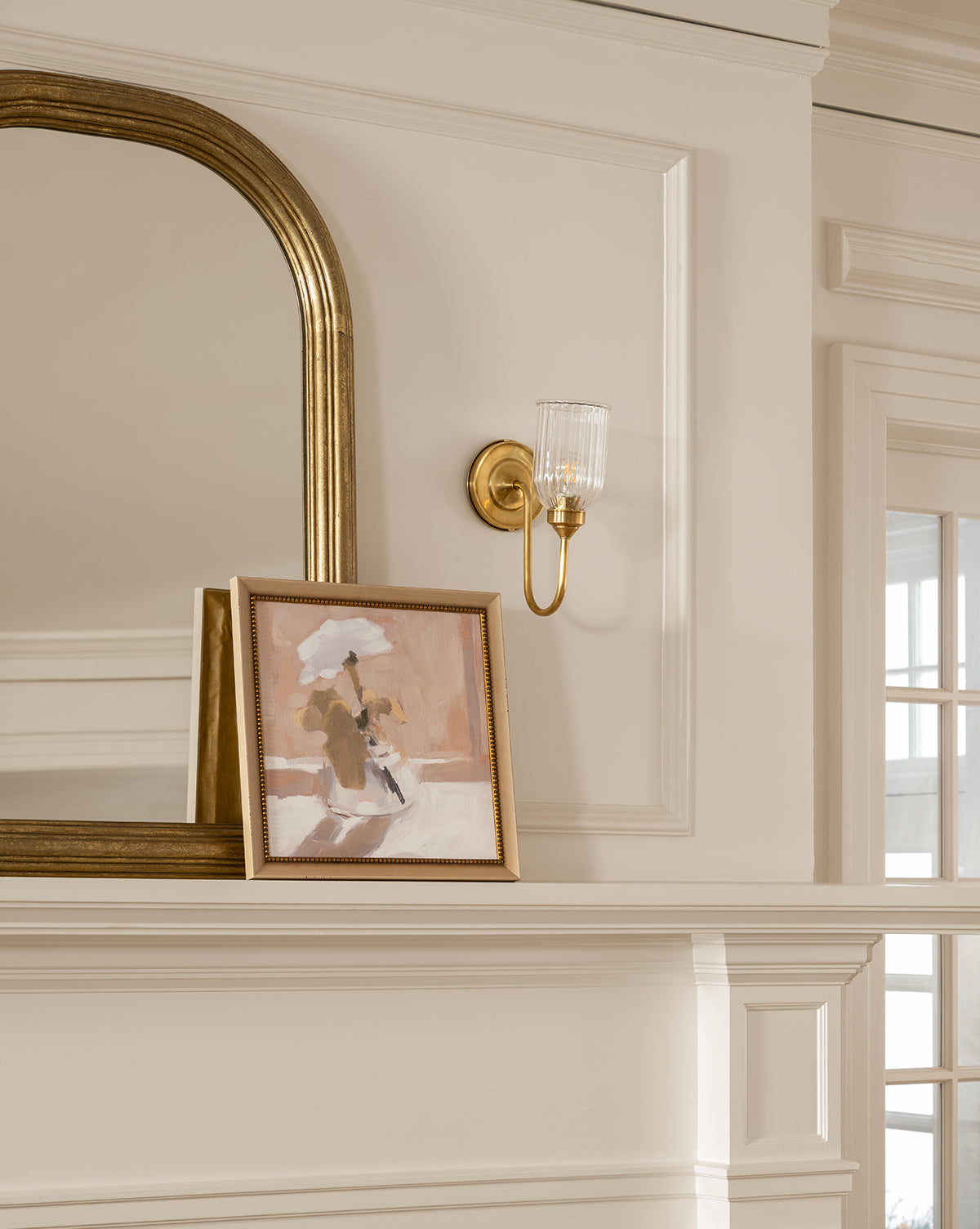 A gold-framed mirror and a gold wall sconce hang above a white mantel. Against the mirror rests Single Blossom, a framed abstract painting, all set within neutral white paneled walls and windows by McGee & Co.