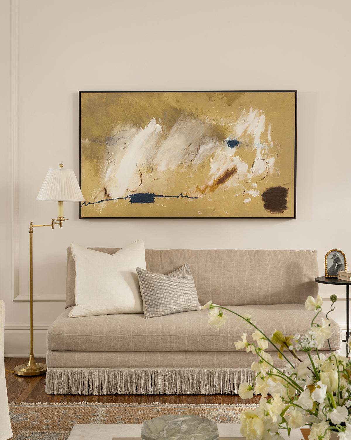 A beige sofa with fringe trim and two pillows sits against a white wall. Above it hangs the Mark Making wall art in yellow, white, blue, and brown. Nearby are a brass floor lamp, a side table with decor, and flowers in the foreground, by McGee & Co.