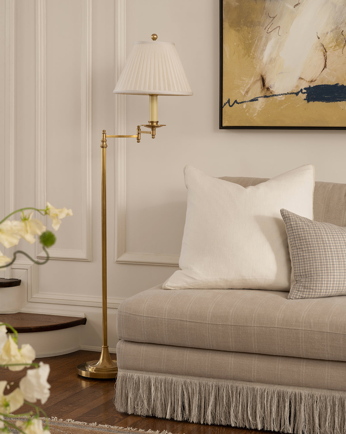 A cozy living room corner with a beige sofa, white and plaid pillows, abstract wall art, wooden stairs in view, and the Dorchester Swing Arm Floor Lamp featuring an antique brass finish and pleated silk shade, by McGee & Co.