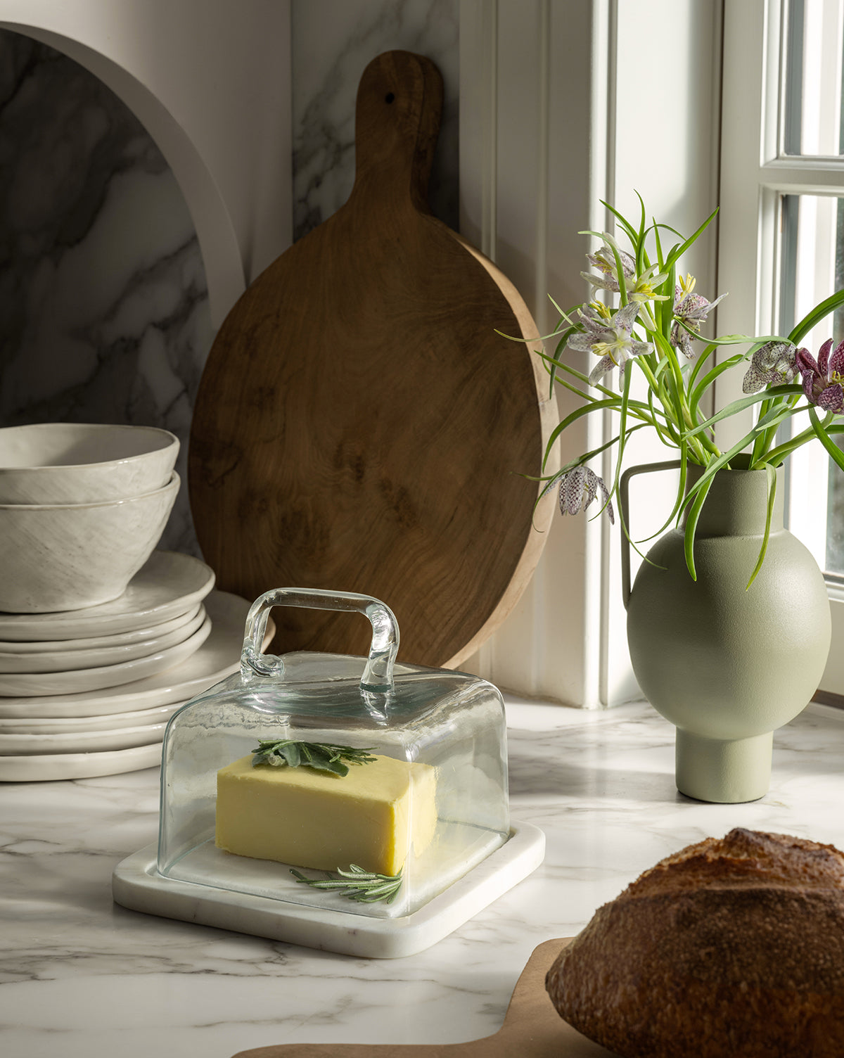 A stick of herbed butter sits on a marble base under the Glass & Marble Cloche, displayed beside stacked white dishes, a wooden cutting board, a green vase with flowers, and a loaf of bread, by McGee & Co.